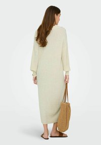 Lång, ribbad stickad klänning i ljusbeige med krage, vida ärmar och längd till mitten av vaden. Modellen håller en vävd brun väska.