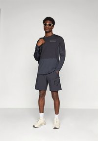 Tamsiai pilka ilgomis rankovėmis marškinėliai su šviesesniu skydeliu ir logotipu, derinami su atitinkančiais šortais su emblemą. Baltos sportbačiai ir saulės akiniai.