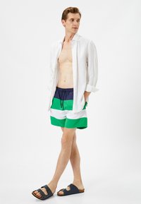 Camisa de lino blanca, pantalones cortos de baño a rayas en azul marino y verde, y sandalias negras. Los pantalones cortos de baño tienen una cintura elástica y cordones.