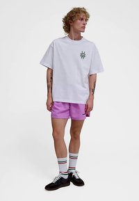 Valkoinen ylisuurikokoinen t-paita, jossa vihreä logo, yhdistetty vaaleanpunaiseen shortseihin. Asusteina raidalliset sukat ja tummat lenkkarit.