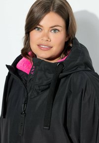 Chaqueta exterior negra con un cuello alto, forro de forro polar rosa, cordones ajustables y herrajes de cremallera. Acabado de tela texturizada.
