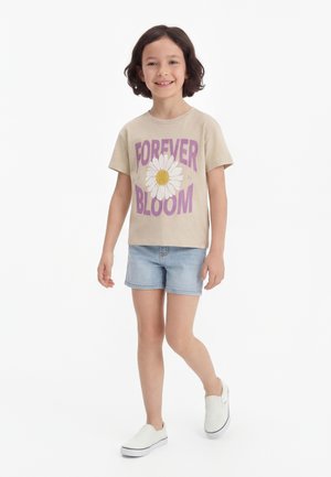 Enfant souriant marchant, portant un T-shirt beige avec le texte « Forever Bloom » et un motif marguerite, un short en jean clair et des chaussures blanches sans lacets.