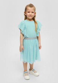 Een lichtblauwe jurk met ruches en doorzichtige mouwen en een sprankelende taille. De rok is van gelaagd tule met kleine sterpatronen. Gecombineerd met witte sneakers.