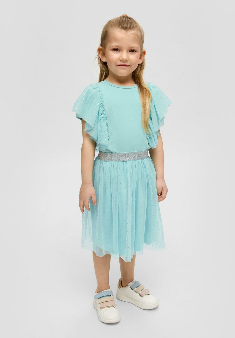 Een lichtblauwe jurk met ruches en doorzichtige mouwen en een sprankelende taille. De rok is van gelaagd tule met kleine sterpatronen. Gecombineerd met witte sneakers.