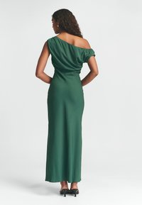 Zelené saténové večerní šaty s asymetrickým výstřihem a drapovaným živůtkem. Dlouhá, přiléhavá sukně až na zem, doplněná sandály na podpatku.