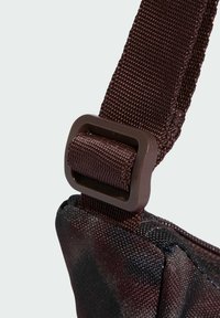 Brązowy pasek nylonowy z plastikową klamrą, z teksturowaną powierzchnią i ciemnym tkaninowym wzorem w miejscu mocowania.