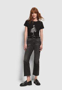 Mujer con cabello rojo vistiendo una camiseta gráfica negra, jeans cortos negros deslavados con dobladillos sin terminar, cinturón negro y mocasines negros, de pie frente a un fondo gris.