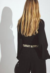 Črn dolg rokav top, kombiniran s črnimi hlačami. Opazen pas s vzorcem kače poudarja pas. Gladki, ravni rjavi lasje padajo dol.