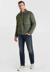 Groene wollen mix jas met rits en kraag, twee zakken aan de voorkant en een gestructureerd oppervlak, gecombineerd met donkerblauwe spijkerbroek en lichte sneakers.