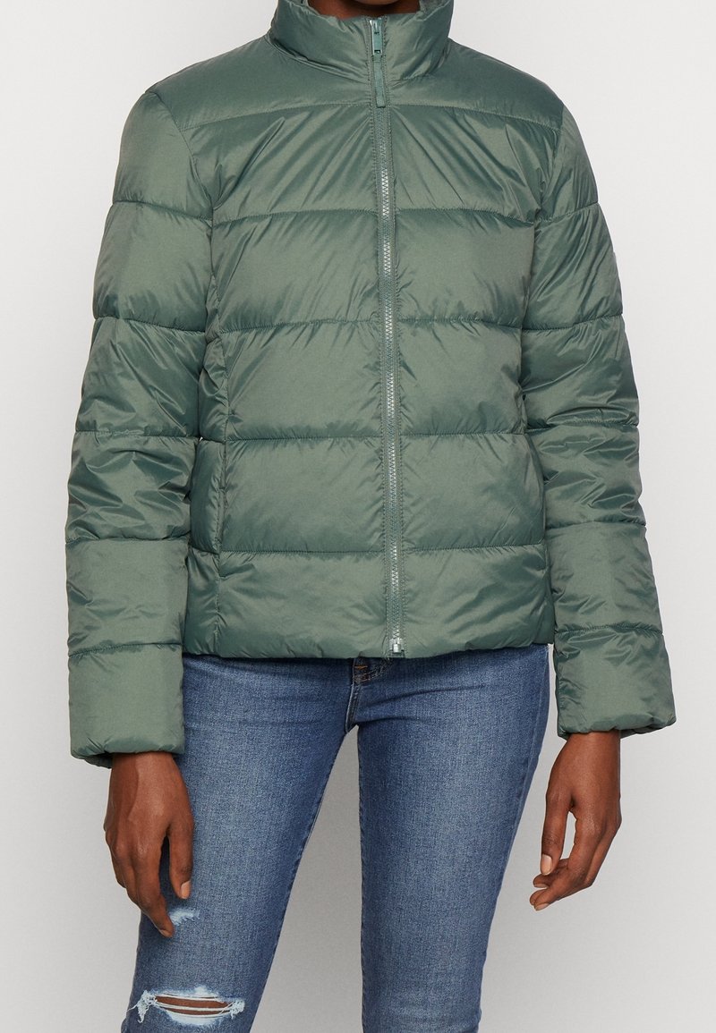 Giubbotto imbottito di un verde tenue, con colletto alto, chiusura con zip e cuciture orizzontali. Indossato con jeans blu strappati. Tessuto morbido.