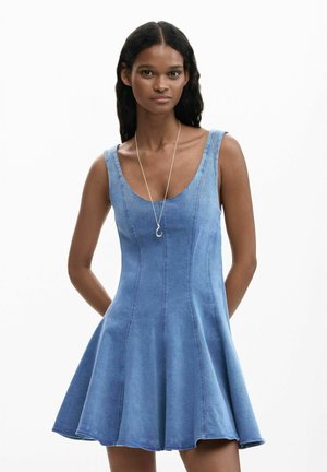 Donna che indossa un vestito di jeans blu senza maniche con gonna svasata e una collana con ciondolo a forma di gancio argentato, in piedi davanti a uno sfondo bianco.