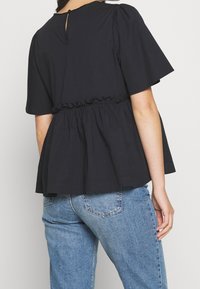 Schwarzes Peplum-Oberteil mit kurzen Ärmeln, gerafftem Taillenbund und einem Schlüssellochverschluss hinten. Weicher Stoff mit glatter Textur. Kombiniert mit hellblauen Jeans.