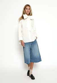 Biela košeľa s veľkou kravatou na golieri, skombinovaná so širokými svetlomodrými denimovými culottes. Vyzerajú skvele s čiernymi loafers a béžovými rebrovanými ponožkami.