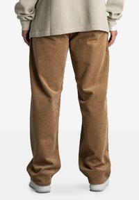 Osoba ubrana w luźne beżowe spodnie kordowe, beżową teksturowaną koszulę z długim rękawem i białe sneakersy, stojąca tyłem do aparatu.