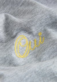 Siva tkanina z mehko teksturo, na kateri je rumeno vezen napis "Oui." Dizajn poudarja kontrast med barvami.