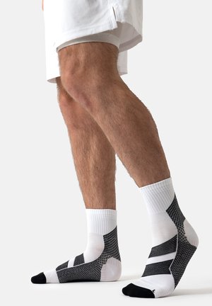 Person wearing white and black athletic ankle socks and white shorts, showing muscular lower legs and feet on a plain background.