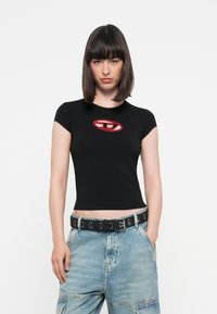Zwarte aansluitende t-shirt met korte mouwen en een rood ovaal logo op de borst, gecombineerd met lichtblauwe wijde jeans en een zwarte gesp riem.