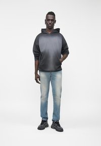 Άνδρας μοντέλο στέκεται φορώντας μαύρο και γκρι φούτερ με κουκούλα, ξεθωριασμένο μπλε τζιν και μαύρα αθλητικά παπούτσια σε απλό λευκό φόντο.