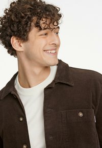 Brun kordflodsskjorta med krage, knappad framtill och bröstficka, lagd över en vit ribbad tröja. Texturerad tyg och knapppdetaljer.