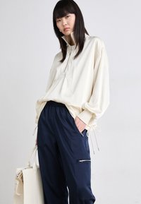 Lys beige genser med halv-lynlås, med en frontlomme og snøring i kanten, kombinert med marineblå bukser med en glidelåslomme og elastisk liv.