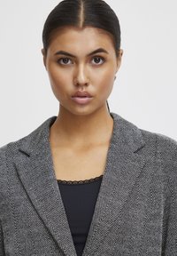 Houndstooth raštuotas švarkas su struktūrizuotu dizainu, pilkos ir juodos spalvų, su notch apykaklėmis ir lygia tekstūra, dėvimas ant juodos palaidinės.