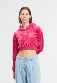 Gekürzter rosa Samt-Hoodie mit einer Kordelzugkapuze, versehen mit einem Logo auf der Brust, kombiniert mit blauen plissierten Jeanshosen.