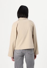 Chaqueta beige, ligera, con mangas largas y cuello con muesca, de corte holgado. Llevada sobre jeans grises.