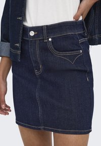 Denim krilo v temno modri barvi, prilegača zasnova s sprednjimi žepi, srebrno gumbno zapiranje in kontrastno šivanje ob šivih ter robu.