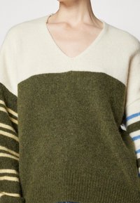 Sweter w serek wykonany z miękkiej dzianiny. Posiada poziome pasy w kolorze oliwkowym i kremowym oraz rękawy w paski w kolorze niebieskim i kremowym.