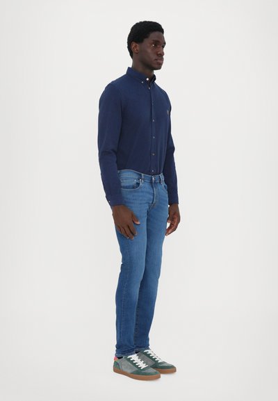 Tengerészeti hosszú ujjú ing, kék slim fit farmernadrág és zöld-szürke velúrbakancs. Az ing gombos gallérral és logó részlettel rendelkezik.