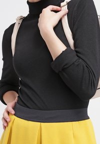 Schwarzer Rollkragenpullover mit enganliegenden Ärmeln, kombiniert mit einem gelben, plissierten Rock mit einem dunklen Taillengummi und beigen Taschenriemen.