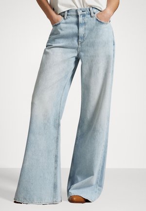 Lichtblauwe, hooggetailleerde wijde jeans, gedragen met handen in de zakken, gecombineerd met bruine schoenen en een wit shirt.