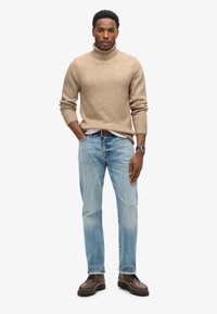 Beżowy sweter z golfem, jasnoniebieskie dżinsy, brązowe buty i zegarek. Posiada teksturowany wzór dzianiny i luźny krój.