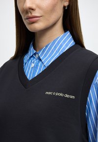 Colete preto de malha com decote em V, usado sobre uma camisa de colarinho azul e branco às riscas. Apresenta o logótipo "marc o polo denim" em dourado.