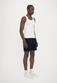 White cotton tank top and navy shorts with drawstring. Model stands in white sneakers and light grey socks against a plain background.