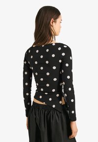 Černý top s dlouhým rukávem s bílými puntíky, má zaoblený lem a boční rozparky. Materiál vypadá pružně a hladce.
