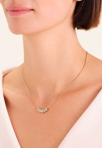 Collana d'oro con cinque diamanti a forma di goccia indossata attorno al collo di una donna su uno sfondo di una maglietta rosa chiaro.