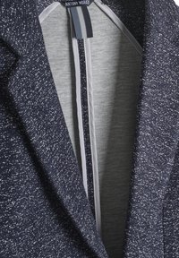 Tamno plavi teksturirani sako sa speckiranim uzorkom; ima sivi oblog i vertikalno zatvaranje na patent zatvarač s prugastim detaljima. Vidljiva oznaka branda.