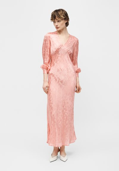 Γυναίκα με μακρύ ροζ σατέν φόρεμα με λουλουδάτο μοτίβο και φουσκωτά μανίκια τριών τετάρτων, συνδυασμένο με λευκά μυτερά παπούτσια.