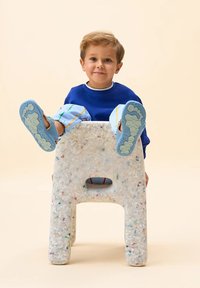 Taburete de plástico reciclado con textura blanca moteada. Un niño se sienta en él, vistiendo suelas de zapatos azules y pantalones a rayas en tonos pastel.