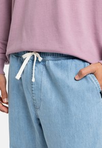 Lyseblå denim joggers med en hvit snor i livet, kombinert med en løse mauve genser. Nærbilde av livet og lommer.