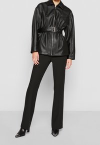 Veste en faux cuir noir avec fermeture éclair, taille ceinturée et accents en métal, associée à un pantalon noir et des bottes à bout pointu.