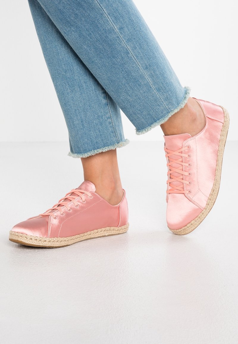 Jambes portant un jean bleu clair à ourlet effiloché et des baskets roses brillantes à lacets avec semelles tissées sur un sol clair.