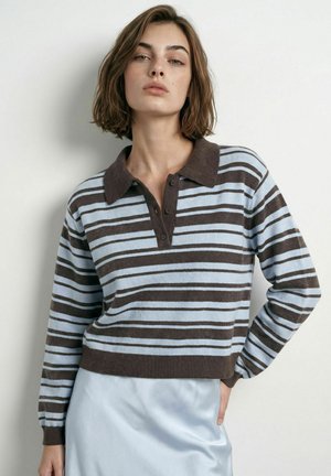 Femme aux cheveux courts et bruns portant un polo à manches longues rayé marron et bleu clair ainsi qu'une jupe en satin bleu clair, debout contre un mur blanc.