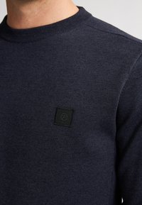 Marineblauwe trui met een ronde hals, gemaakt van gestructureerde stof, met een zwart geweven label op het linkerborstgebied.