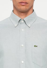 Light grey button-up shirt with a pointed collar, white buttons, and a chest pocket featuring a green crocodile logo.
