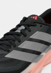 Scarpa da ginnastica nera con tomaia testurizzata, caratterizzata da tre strisce grigie, lacci neri e un accento rosa vicino al tallone. Suola semi-trasparente.