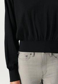 Black long-sleeve top with a gathered waistband, paired with light gray fitted jeans featuring subtle stitching and front pockets.