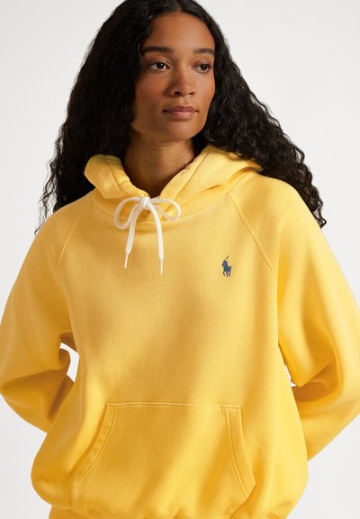 Rumena majica s kapuco iz mehke tkanine. Ima kengurujski žep in bel trak za zategovanje kape. Majhen modri logotip na prsih.