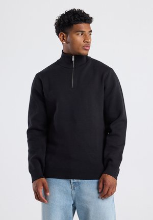 Pull en tricot noir avec un col zippé haut et des poignets côtelés, associé à un jean en denim clair. Texture lisse et design à coupe classique.